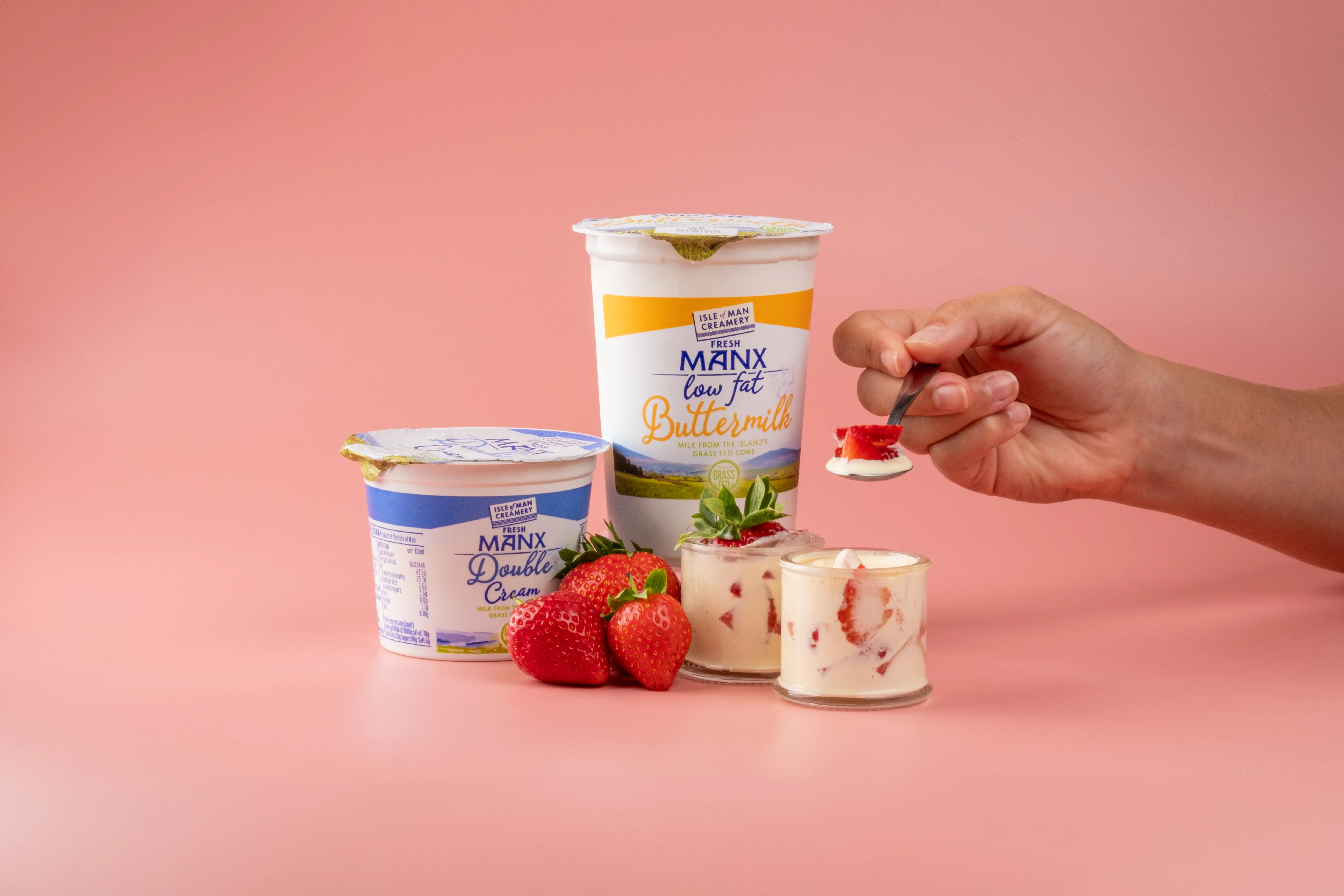 Hands spooning Manx Creamery buttermilk and double cream products styled with fresh strawberries on a pink background